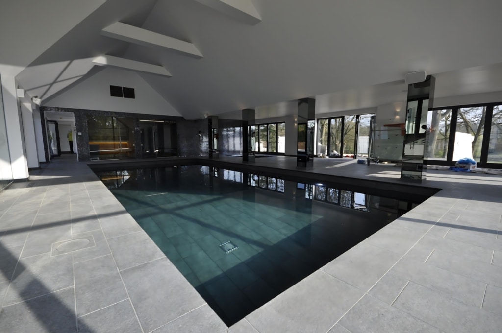 An indoor pool with dark water, surrounded by a gray stone deck, featuring large floor-to-ceiling windows and a modern, high-ceiling design