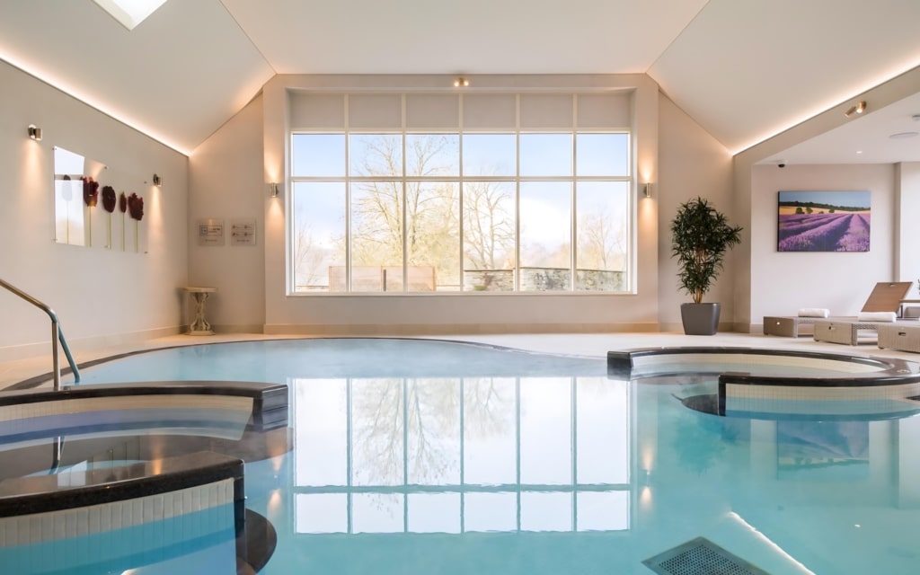 An indoor pool area with multiple interconnected pools, surrounded by a stone deck, featuring large windows, modern deco