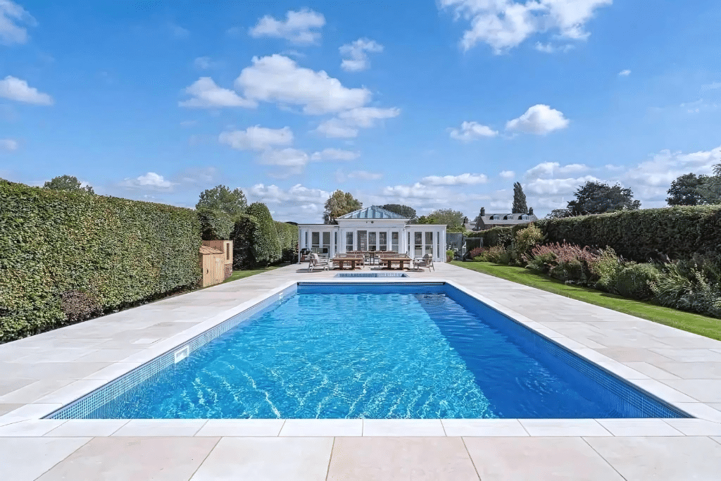 A rectangular swimming pool with blue water, surrounded by a limestone deck, in a landscaped garden with hedges and a white pool house in the background.